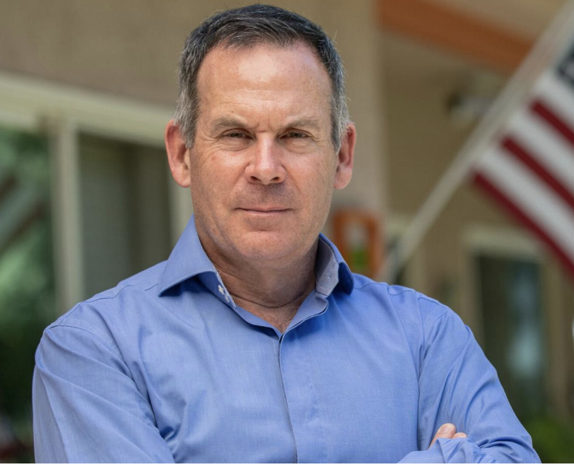 Adam Frisch wearing a light blue collared button up shirt crossing his arms and gazing into the camera.